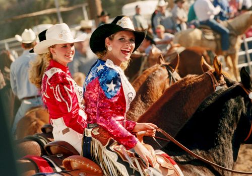 Two women in colorful outfits and cowboy hats are riding horses at a rodeo event, with people and horses in the background.