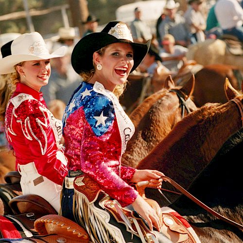 Two women in colorful outfits and cowboy hats are riding horses at a rodeo event, with people and horses in the background.