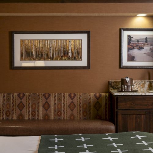 A cozy hotel room features a bed, patterned couch, framed pictures, a lamp, and a small kitchenette area with a sink, coffee maker, and condiments.