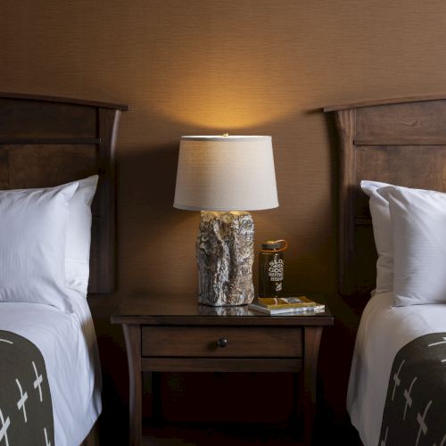 The image shows a hotel room with two beds, a nightstand between them, a table lamp, and decorative pillows on each bed.