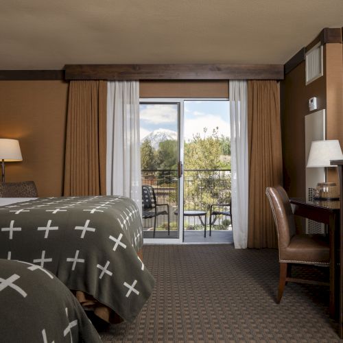 The image shows a hotel room with two beds, a desk with a mirror, a lamp, a chair, and a balcony with a view of trees outside.