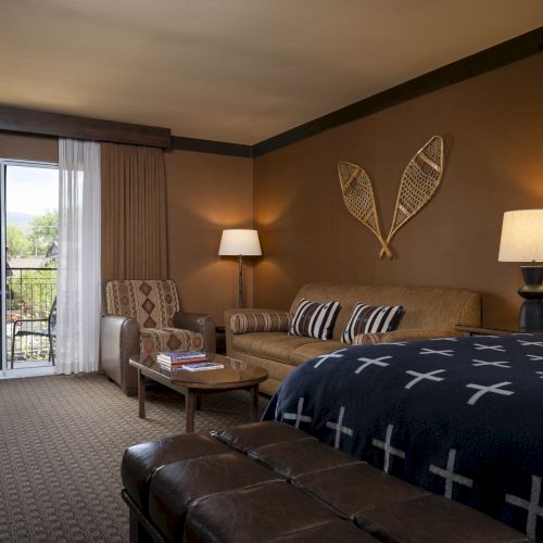 A hotel room with a bed, couch, coffee table, two lamps, and a sliding glass door leading to a balcony is shown.