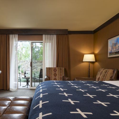 This image shows a hotel room with a large bed, two side tables with lamps, an armchair, a desk, and a sliding door leading to an outdoor area.