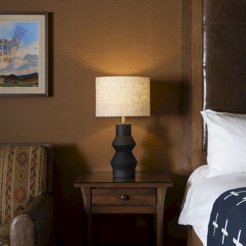 A cozy bedroom with a patterned chair, a wooden nightstand, a modern lamp, a bed with a cross-patterned blanket, and a framed picture on the wall.