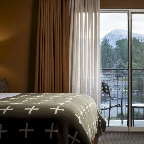 A cozy hotel room with a bed, bedside lamp, and curtains partially open to reveal a balcony view of mountains and trees in the background.