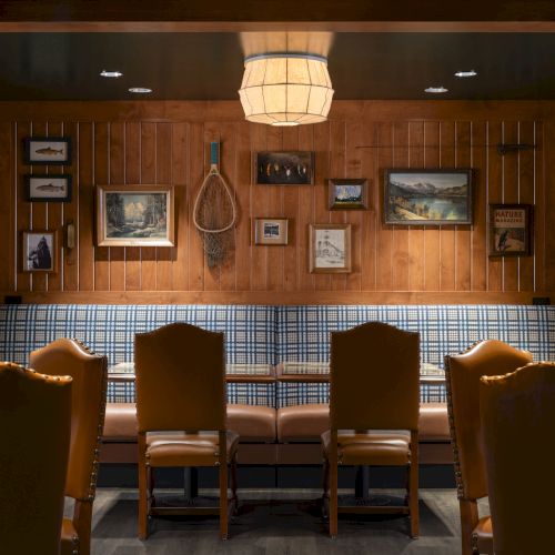 This image shows a cozy nook with wooden paneling, framed pictures, a tennis racket, stone walls, a plaid bench, and leather chairs under soft lighting.