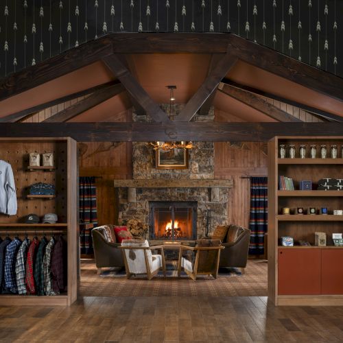 A cozy room featuring a stone fireplace, shelves with clothing and mugs, and a sitting area with armchairs and a table in front of the fire.