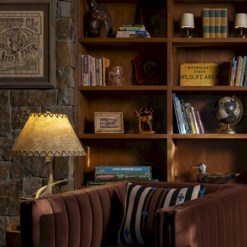 The image shows a cozy room with a bookshelf filled with books and decor, a stone wall, a framed artwork, a lamp, and a cushioned chair.