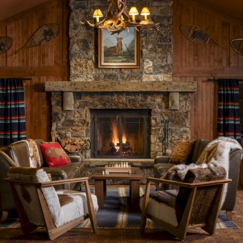 A cozy rustic living room with a stone fireplace, comfortable seating, a chessboard, and warm lighting. Snowshoes and painting decorate the wall.