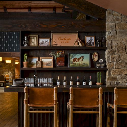 Three bar stools at a wooden bar with shelves displaying photographs, bottles, and decor in a rustic setting.