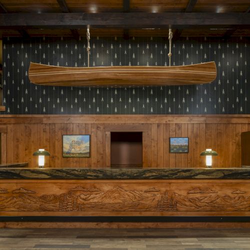 A wooden reception desk with intricate carvings, two lamps, framed pictures, and a suspended canoe in a rustic-themed interior ends the sentence.