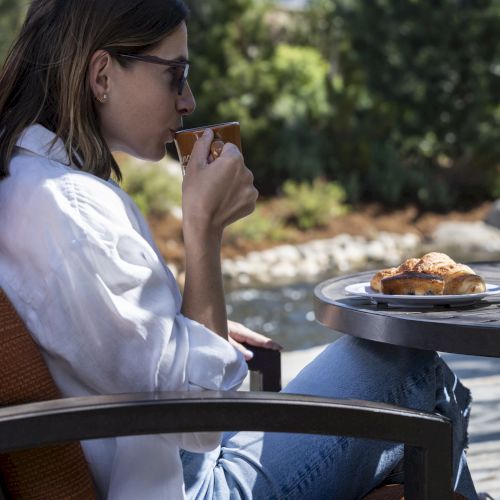 A woman in a white shirt and jeans enjoys a cup of coffee while sitting outdoors, with a plate of pastries on a small table.