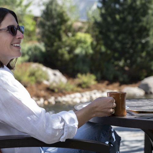 A person is sitting outdoors on a patio chair, wearing sunglasses, and holding a cup while smiling and relaxing.