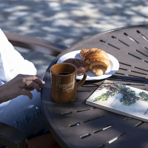 A person is sitting by a table with a cup of coffee, a croissant on a plate, and a book or magazine, enjoying a sunny, shaded outdoor setting.