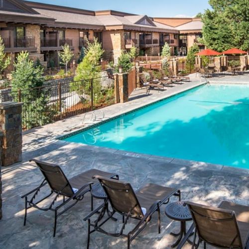 The image shows a well-kept outdoor swimming pool surrounded by lounge chairs, trees, and a fence, within a residential or hotel complex.