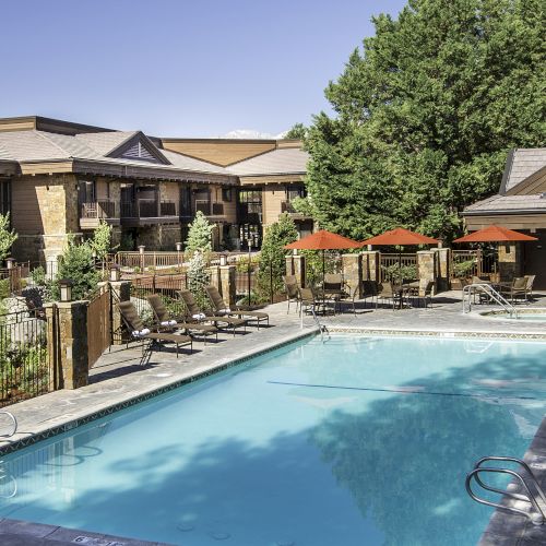 An outdoor swimming pool surrounded by lounge chairs, umbrellas, and a building with balconies and trees.