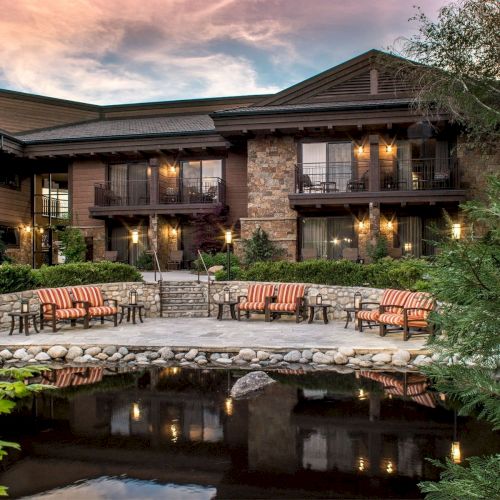 A serene lodge with outdoor seating, surrounded by lush greenery, and overlooking a peaceful pond under a twilight sky.