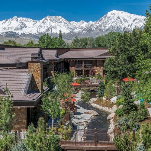 A scenic resort with modern buildings, lush greenery, and outdoor seating areas, surrounded by majestic snow-capped mountains under a clear blue sky.