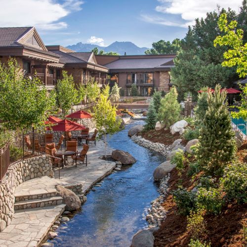 A serene outdoor scene with a stream running through a well-landscaped area, surrounded by buildings, trees, and red patio umbrellas under a blue sky.