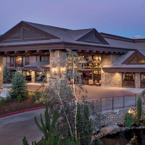 A welcoming lodge with stone and wood elements, surrounded by landscaped greenery and mountains in the background. It looks serene and inviting.