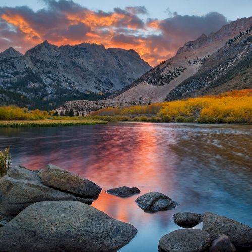 A serene mountain landscape features a calm lake reflecting a vibrant sunset, rocky shoreline, and trees with autumn foliage, set against towering peaks.