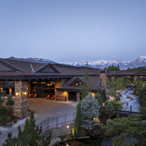 A scenic view of a large lodge-style building with mountains in the background, surrounded by trees and a stream in the foreground.