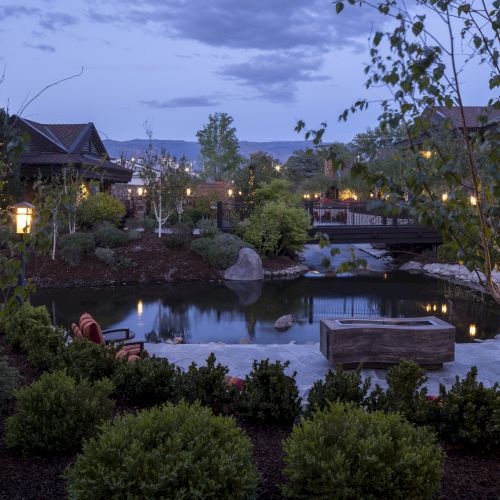 A tranquil outdoor setting with a pond, lush greenery, trees, and softly lit buildings at dusk, creating a serene atmosphere.