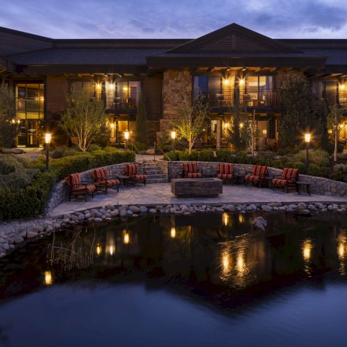 The image shows a luxurious building with outdoor seating around a fire pit, beautifully illuminated at night, reflected in a calm pond.