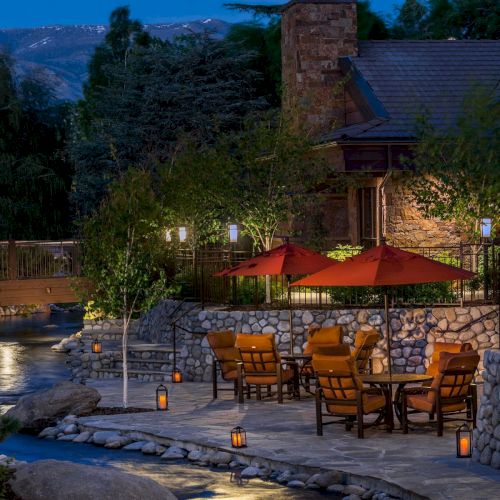 An outdoor patio with seating, red umbrellas, lanterns, and stone walls next to a stream in a scenic, wooded area during dusk.