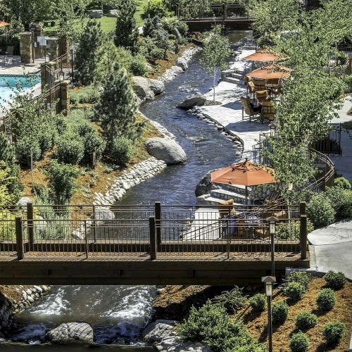 An outdoor area with a pool, a river spanned by bridges, walking paths, trees, and seating areas with orange umbrellas situated by the river.