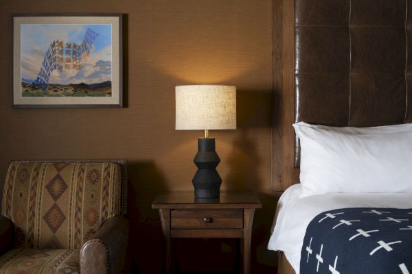 The image shows a cozy bedroom with a patterned armchair, a bedside table with a lamp, and part of a bed with a decorative blanket.