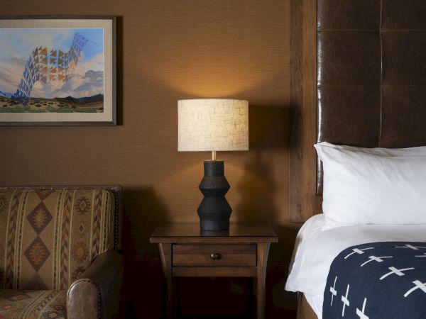 The image shows a cozy bedroom with a patterned armchair, a bedside table with a lamp, and part of a bed with a decorative blanket.