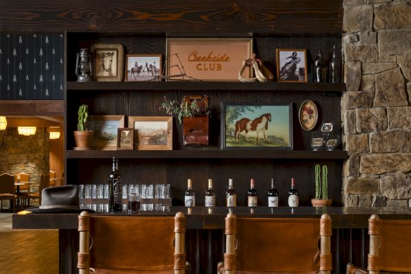 The image features a cozy bar area with wooden stools, framed pictures, and bottles on display, complemented by stone walls and warm, rustic decor.