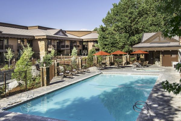 An outdoor swimming pool at a residential complex with lounge chairs, red umbrellas, and adjacent greenery under a clear sky.