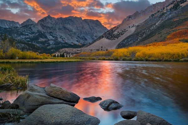 A serene lake reflects colorful clouds and rugged mountains at sunset. Yellow foliage lines the shore, with rocks in the foreground.