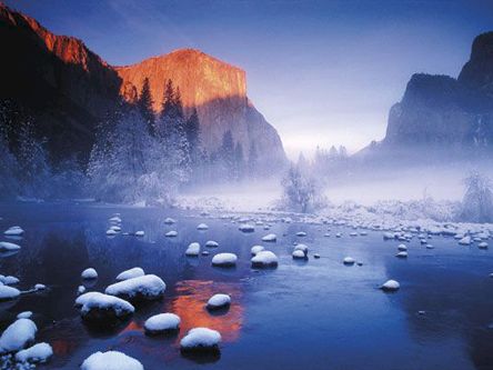 A misty winter lake with snow-covered rocks, floating ice, and towering mountains under a glowing sunrise.