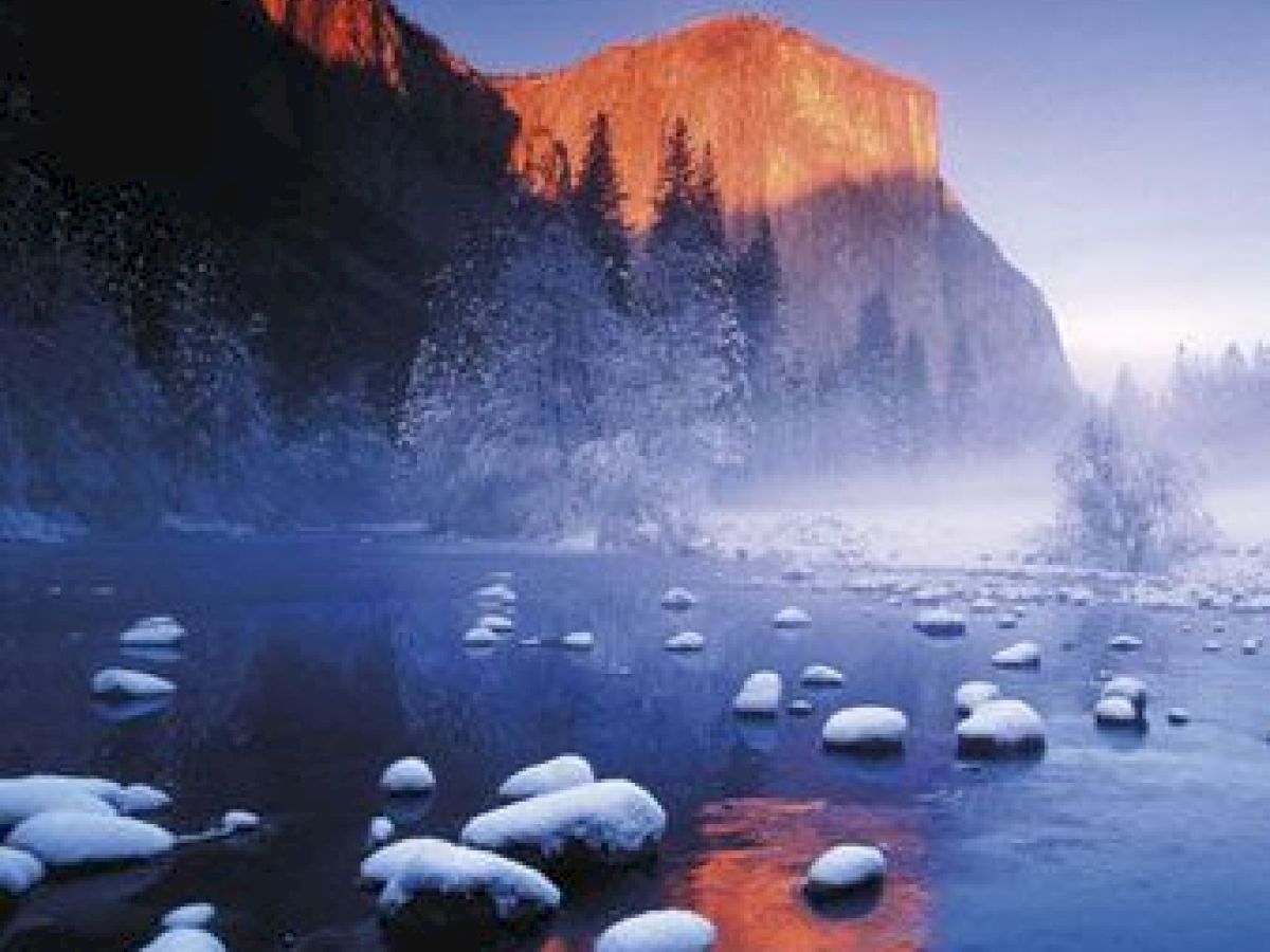 The image depicts a misty landscape with a river, snow-covered rocks, trees, and a sunlit mountain peak in the background.