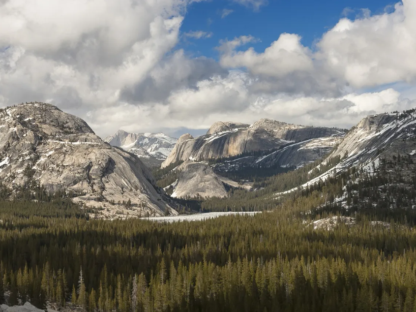 A vast mountain valley with snowy peaks, dense autumn forests, and dramatic clouds under a bright blue sky.