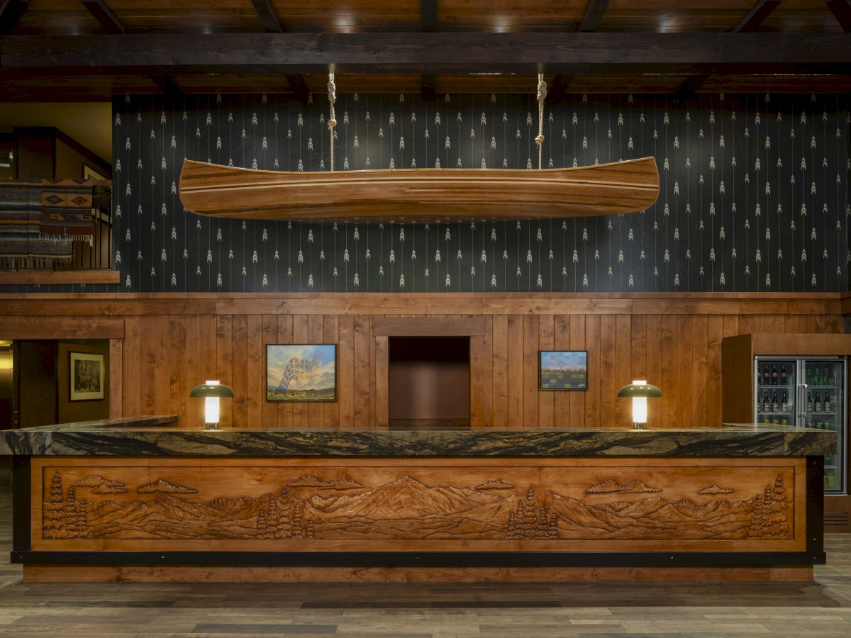 A hotel lobby shows a large hanging wooden canoe over a carved reception desk with patterned curtains and warm lighting.