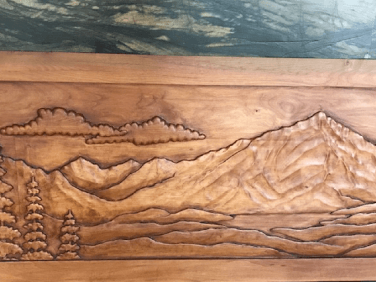 The image shows a wood carving depicting a mountainous landscape with trees and clouds in the background.