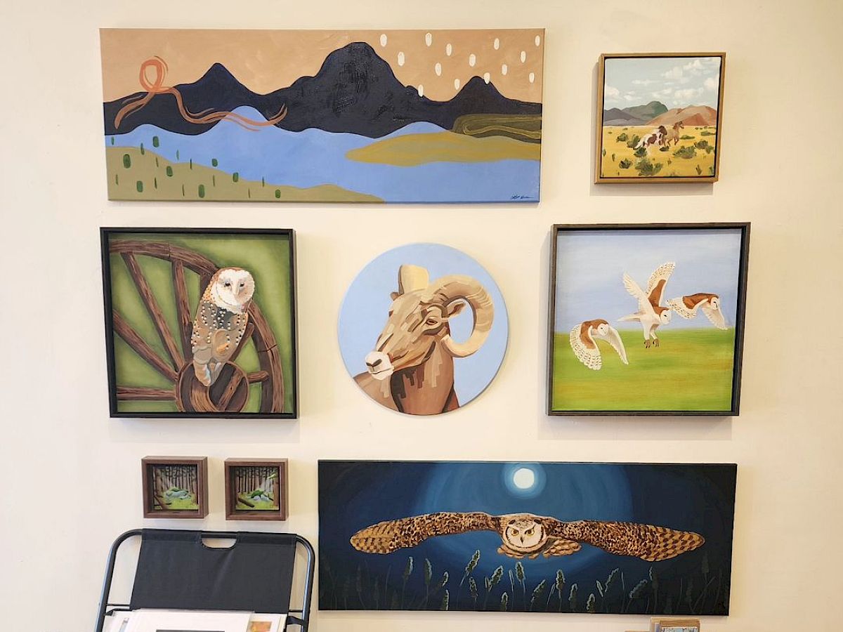 The image shows a wall gallery with various animal and landscape paintings, a small table with items, and a black rack holding papers or prints.