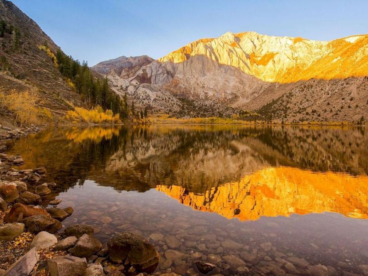 The image shows a serene mountain landscape with rocky slopes, trees, and a clear lake reflecting the mountains and sky at sunset.