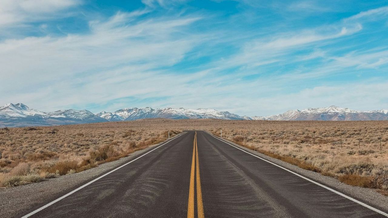 A straight road stretches into the distance through a barren landscape, under a blue sky dotted with clouds, with mountains on the horizon.