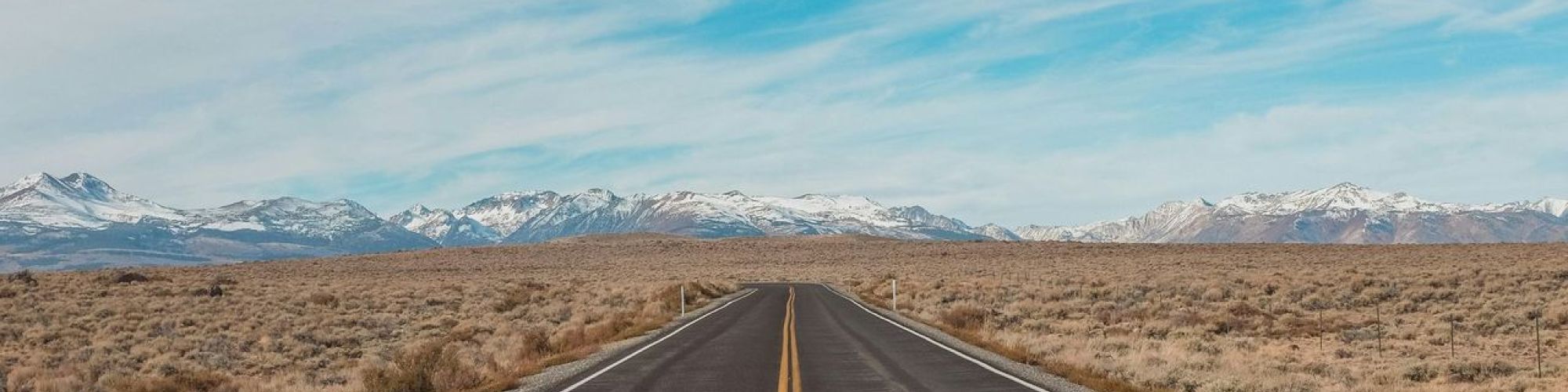 A straight road stretches into the distance through a barren landscape, under a blue sky dotted with clouds, with mountains on the horizon.