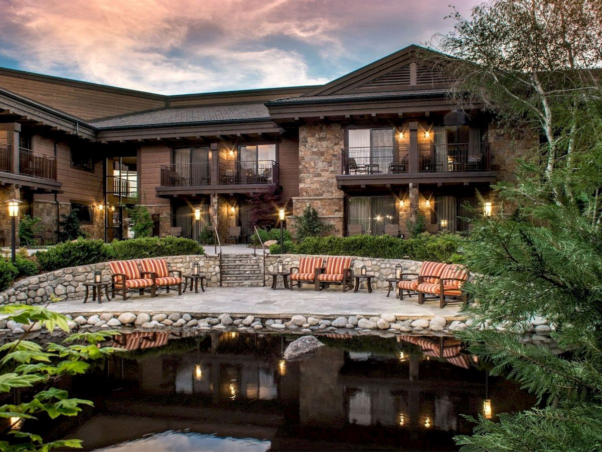 A tranquil outdoor area with lounge chairs is seen in front of a rustic building surrounded by lush greenery and a serene pond.