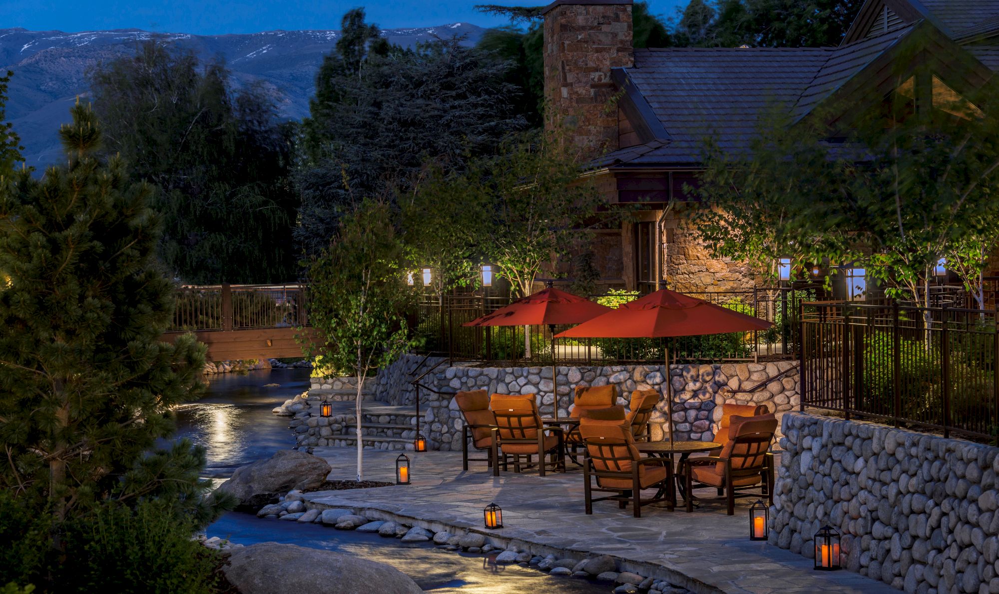 An outdoor patio with red umbrellas, cushioned chairs, stone pavement, trees, and hanging lanterns, all set beside a small stream near a forested area.