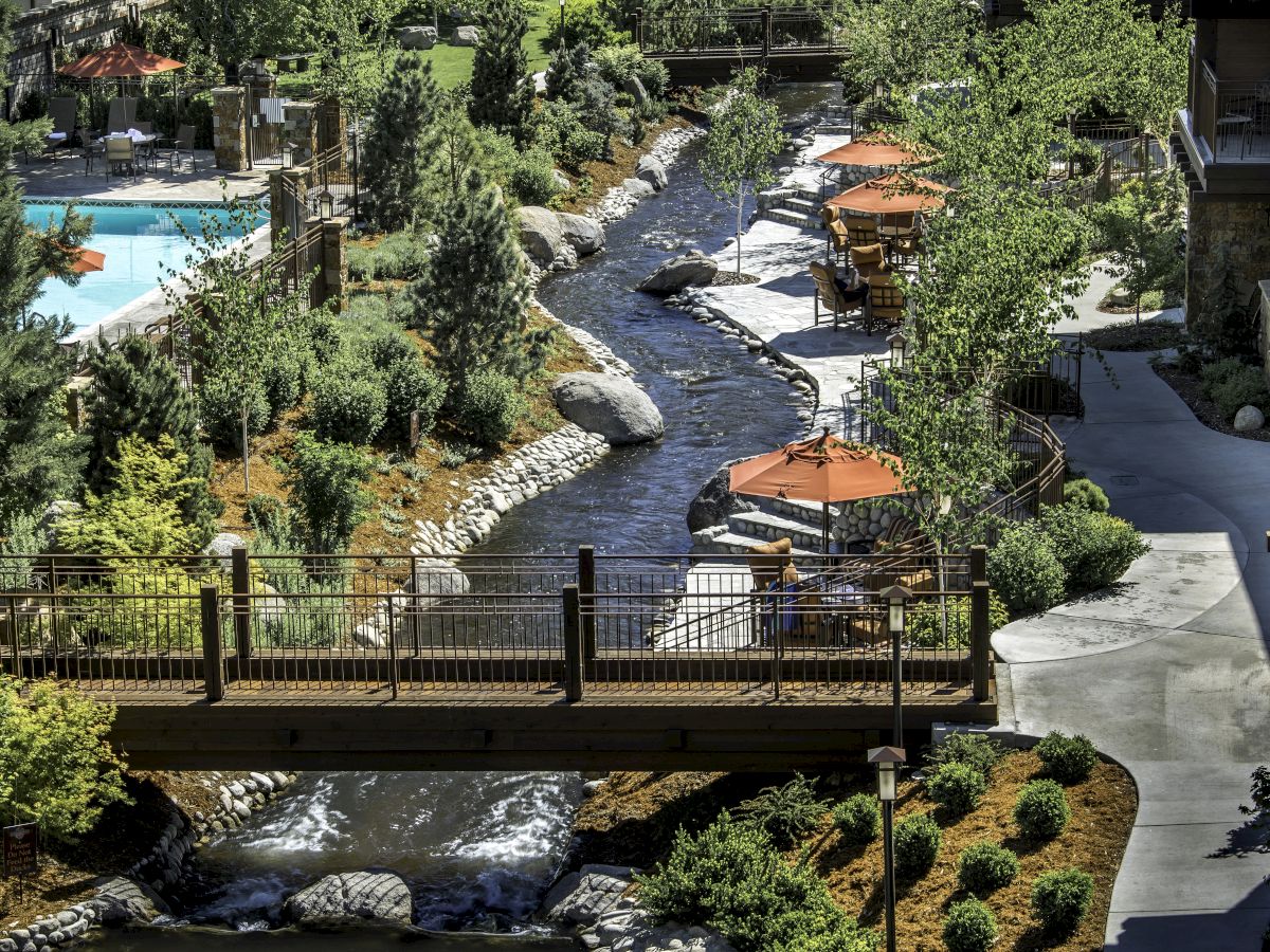 A scenic view of a resort with a bridge over a stream, outdoor seating with red umbrellas, lush greenery, and a pool area to the left.