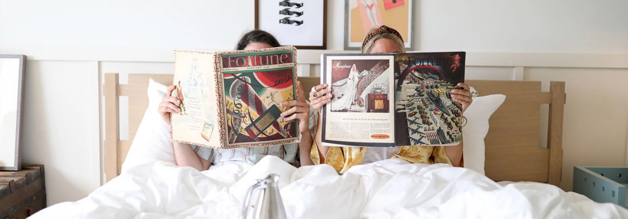 Two people are lying in bed, covered with a white blanket, reading magazines, with framed art visible on the wall behind them.