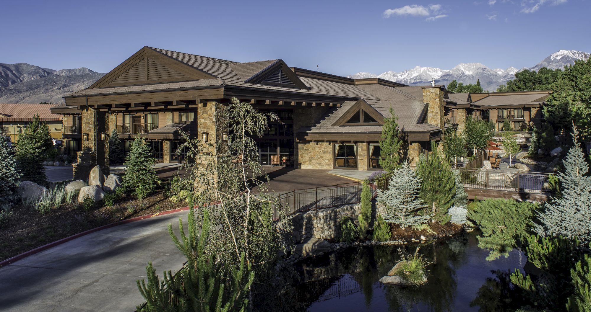 A large, upscale lodge-style building with stone and wood accents, surrounded by trees and a small pond, set against mountains in the background.