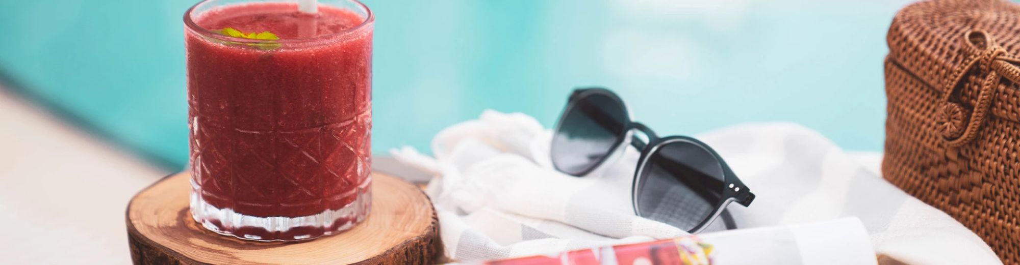 A red drink in a glass on a wooden coaster, sunglasses, a woven bag, and a rolled-up towel by a poolside setting.